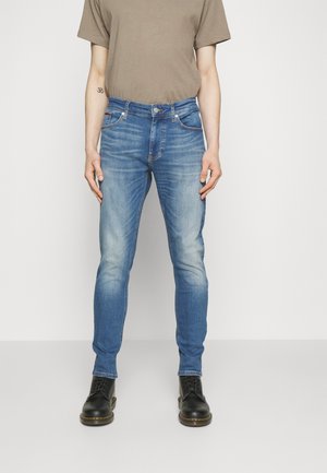 Jeans slim-fit azzurri chiari con sbiaditure, cinque tasche e chiusura a bottone. Indossati con una T-shirt semplice color oliva e stivaletti neri.
