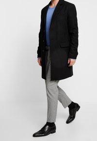 Cappotto di lana nero con maniche lunghe, design a un solo bottone, due tasche frontali, abbinato a un maglione blu e pantaloni a quadri. Scarpa in pelle nera.