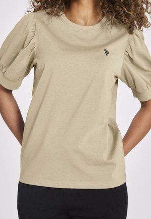 Woman wearing a beige puff-sleeve shirt with a small black embroidered logo on the left chest, paired with black pants.