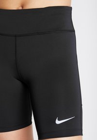 Detailní záběr černých sportovních trenýrek Nike nošených v pase, ukazující hladkou látku, ploché švy a bílý logo swoosh Nike na stehně.