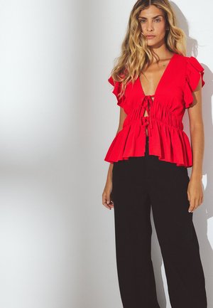 Červený peplum top s rozšírenými rukávmi a hlbokým V-výstrihom, s viazaním. V páre s čiernymi širokými nohavicami.