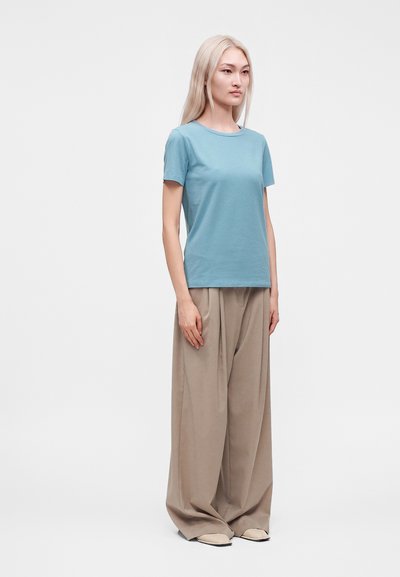 Femme aux longs cheveux blonds portant une chemise bleu clair à manches courtes et un pantalon large beige, debout devant un fond uni.