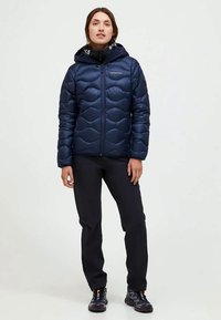 Woman wearing a navy quilted jacket with hood, black pants, and black athletic shoes, standing against a plain white background.