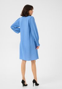 Robe en coton bleu avec une surface texturée, manches longues et détails smockés aux poignets. Mi-longue et coupe ample. Portée avec des escarpins noirs.