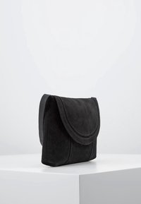 Černá semišová taška přes tělo s klopou, odpočívající na bílé podstavě na pozadí s jednoduchým světlým pozadím.