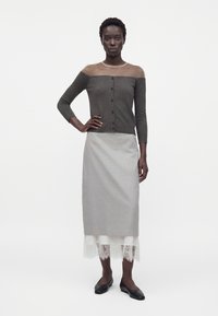 Donna che indossa un cardigan grigio con spalle scoperte e bottoni, una gonna midi grigia con orlo in pizzo bianco e ballerine nere, in piedi con una mano sul fianco.