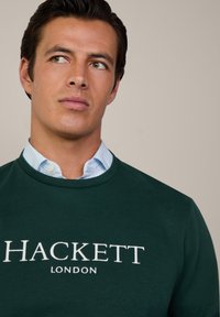 Tmavohnedý mikina s bielym logom "HACKETT LONDON". Prehodená cez svetlomodrú a bielo pruhovanú košeľu s golierom. Hladká textúra.