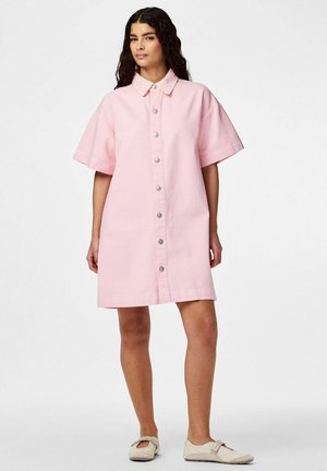 Vrouw met lang donker haar die een losse, korte mouwen, roze jurk met knopen draagt, gecombineerd met lichtbeige platte schoenen, staand op een witte achtergrond.