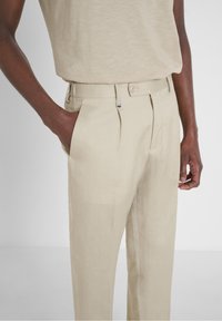 Muž ve světlých lněných kalhotách a odpovídající béžové košili, s jednou rukou v kapse, před jednobarevným světlým pozadím.