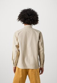 Light beige long-sleeve shirt in cotton with a button-down collar, featuring side seam stitching and buttoned cuffs, worn with yellow shorts.