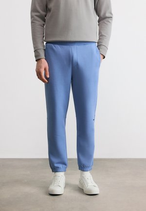 Homme portant un pantalon de jogging bleu clair, un sweat-shirt gris et des baskets blanches, debout sur un sol gris contre un mur blanc.