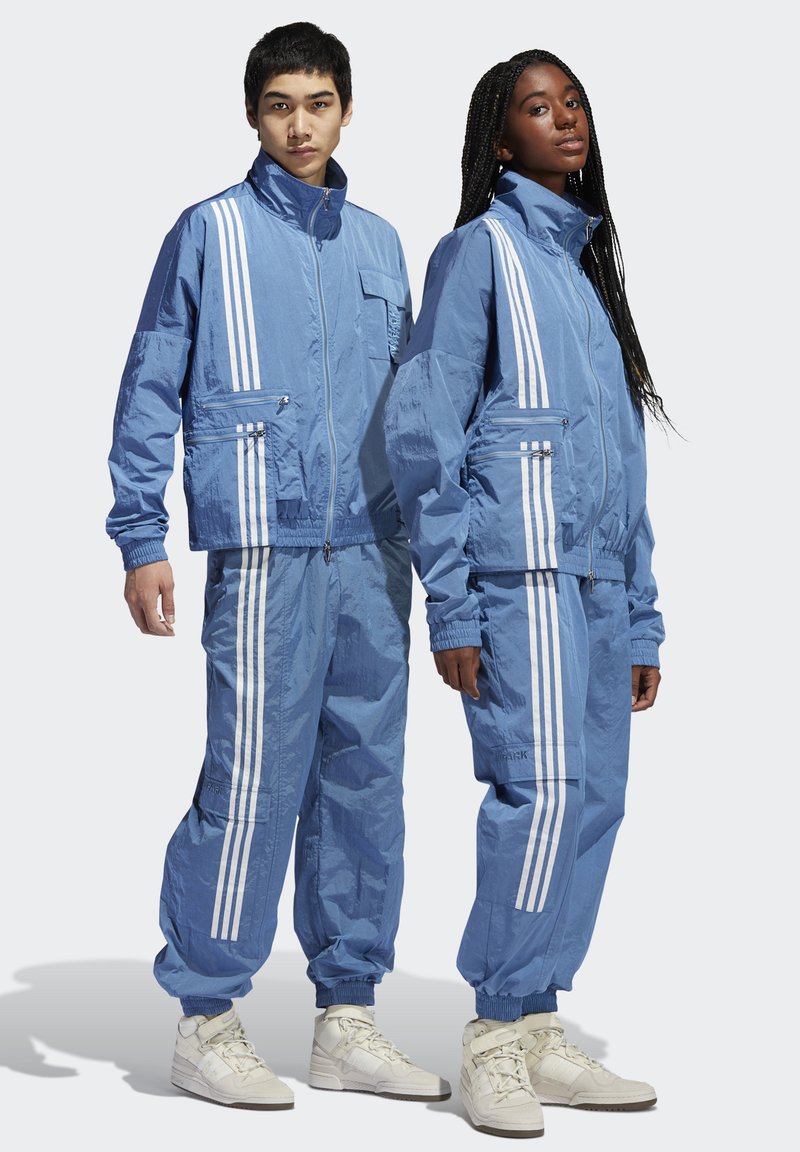 Twee modellen in bijpassende blauwe sportieve trainingspakken met witte strepen en witte hoge sneakers, staan zij aan zij.