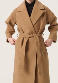 Brauner Wollmantel mit Taillengürtel, weitem Kragen und langen Ärmeln. Verfügt über Seitentaschen und eine weiche Textur.