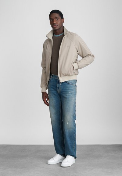 Μπεζ σακάκι με διπλωμένο γιακά, υφασμάτινη καφέ μπλούζα από κάτω, ανοιχτό μπλε τζιν με ξεθωριασμένες λεπτομέρειες και λευκά αθλητικά παπούτσια.