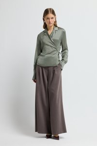 Hellgrünes Satin-Wickelshirt mit seitlicher Schleife, kombiniert mit locker sitzenden, dunkelgrauen Weitbein-Hosen und tiefburgunderroten spitzen Schuhen.