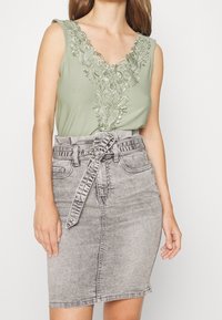 Haut sans manches vert avec des broderies décoratives autour de l'encolure, associé à une jupes en denim grise dotée d'une taille ceinturée et d'un zip sur le devant.