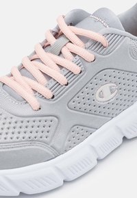 Baskets de sport gris clair avec des lacets roses, tige en maille avec des détails perforés, et semelle extérieure en caoutchouc blanc avec des rainures texturées.