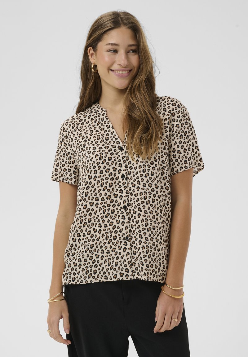 Femme souriante aux longs cheveux portant un chemisier à boutons imprimé léopard et un pantalon noir, accessoirisée avec des boucles d'oreilles et des bracelets en or.