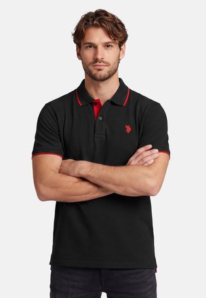 Mann mit hellbraunen Haaren und Bart, der ein schwarzes Poloshirt mit rotem Besatz trägt, steht mit verschränkten Armen vor weißem Hintergrund.