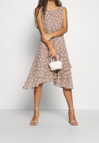 Braunes Polka-Dot-Midi-Kleid mit ausgestelltem Saum und geschichtetem Design, kombiniert mit einer weißen Mini-Handtasche und beigen Blockabsatzsandalen.