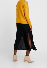 Geel trui met geribde zoom, gecombineerd met een zwarte geplooide rok met een zijsplit, en beige enkellaarsjes met een lage hak.