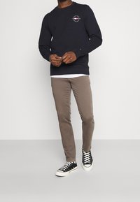Granatowy sweatshirt z logo na klatce piersiowej, zestawiony z jasnobrązowymi spodniami slim-fit i czarnymi niskimi trampkami z białymi podeszwami i sznurowadłami.