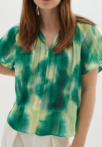 Blusa a maniche corte, verde e gialla con un motivo a fantasia, con scollatura a V e vestibilità comoda. Realizzata in tessuto leggero e trasparente con una texture liscia.