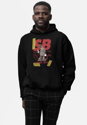 Mężczyzna ubrany w czarną bluzę z kapturem z nadrukiem kreskówkowego głośnika jadącego na deskorolce oraz czerwonym tekstem, w połączeniu z czarnymi spodniami z białą kratką.