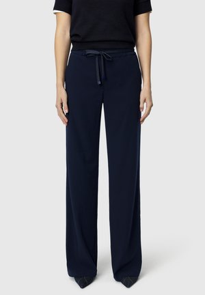 Mujer vestida con pantalones anchos de pierna en azul marino con cintura anudada y camiseta negra, posando contra un fondo claro y sencillo.