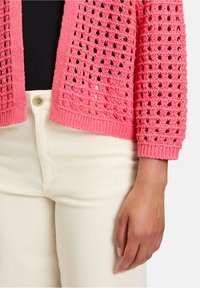 Cardigan en maille ouverte de couleur rose vif, avec un tissage lâche, des poignets à côtes et un ourlet à côtes. Assorti à un jean couleur crème.