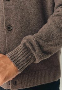 Brown knitted cardigan with ribbed cuffs, featuring a soft texture and dark buttons. The sleeve is slightly rolled at the wrist.