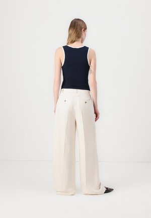 Vrouw met een mouwloze marineblauwe top en hoge taille, wijd uitlopende witte broek met fijne strepen, staand met haar rug naar de camera tegen een effen achtergrond.