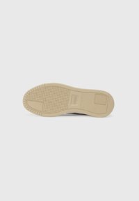 Sneaker sole in light beige rubber with textured patterns; includes a rectangular branding patch. Shaped for grip and traction.