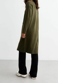 Cardigan lungo a maglia verde oliva con orlo a coste, due tasche e vestibilità morbida, abbinato a pantaloni neri e scarpe da ginnastica bianche.