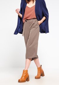 Culotte marroni con cintura annodata, abbinati a una canotta ruggine e a un blazer blu con mantella. Indossati con stivaletti con tacco color beige.