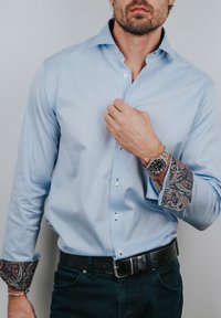 Man die een lichtblauw overhemd met paisley-patroon aan de manchetten verstelt, draagt een zwarte riem, donkere spijkerbroek en een zilveren polshorloge.