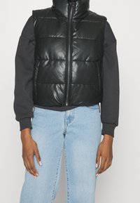 Svart vadderad väst med dragkedja och hög krage, tillverkad av glänsande material, lager ovanpå en grå sweatshirt, kombinerad med ljusblå jeans.