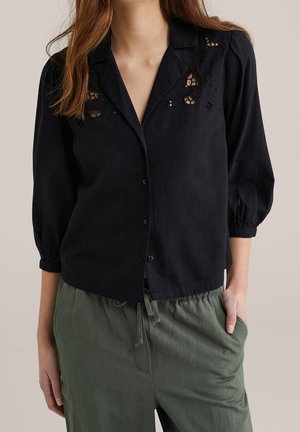 Femme portant une chemise noire boutonnée avec des découpes florales près du col et un pantalon vert ample avec une taille à cordon, main dans la poche.