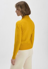 Geel coltrui met een aansluitend ontwerp, geribbelde zoom en ruches bij de halslijn, gecombineerd met crèmekleurige broek.