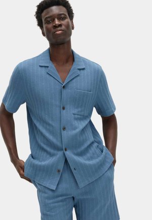 Mand iført en blå kortærmet skjorte med knapper og matchende shorts med hænderne i lommerne foran en ensfarvet baggrund.