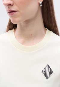 Κρεμ φούτερ με στρογγυλή λαιμόκοψη, διαθέτει μαύρο διαμάντι κεντημένο με κάθετες ρίγες στο μπροστινό μέρος.