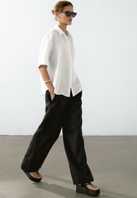 Camicia bianca in lino a maniche corte, pantaloni neri a gamba larga e sandali con piattaforma neri. Gli accessori includono un bracciale in argento e occhiali da sole.