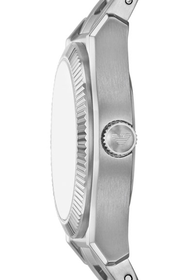 Silverfärgad klocka i rostfritt stål med en texturerad bezel och rund klockkapsel. Har en framträdande krona på sidan, utan synliga detaljer på urtavlan.