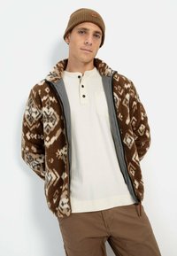Bruine fleecejack met geometrische patronen, grijze accenten, ritssluiting en een kraag. Draag het over een lichtgekleurde henley shirt en bruine broek.