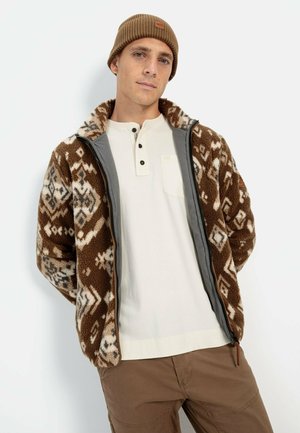 Braune Fleecejacke mit geometrischen Mustern, grauen Akzenten, Reißverschluss und Kragen. Getragen über einem hellen Henley-Shirt und braunen Hosen.