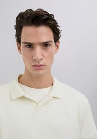 Tricou polo galben pal cu țesătură texturată și guler clasic, purtat peste un tricou crem deschis. Fundal simplu.