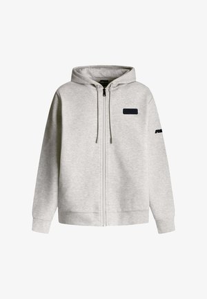 Grå zip-up hoodie med huva, tillverkad av mjukt material. Har en liten mörk fläck på bröstet och en mörk detalj på vänster ärm.