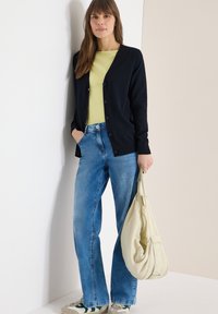 Femme portant un cardigan bleu marine, un haut vert citron, un jean bleu et des baskets, tenant un sac à main souple beige, debout devant un mur bicolore.