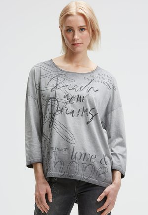 Blonde Frau, die ein lockeres graues Langarmoberteil mit motivierendem Text, einschließlich "Erreiche deine Träume", und verblasste dunkelblaue Jeans trägt.