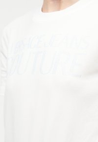 Λευκό πουκάμισο με αχνό, φωτεινό μπλε κείμενο από στρας που γράφει «Versace Jeans Couture» στο πάνω μέρος του στήθους ενός ατόμου.